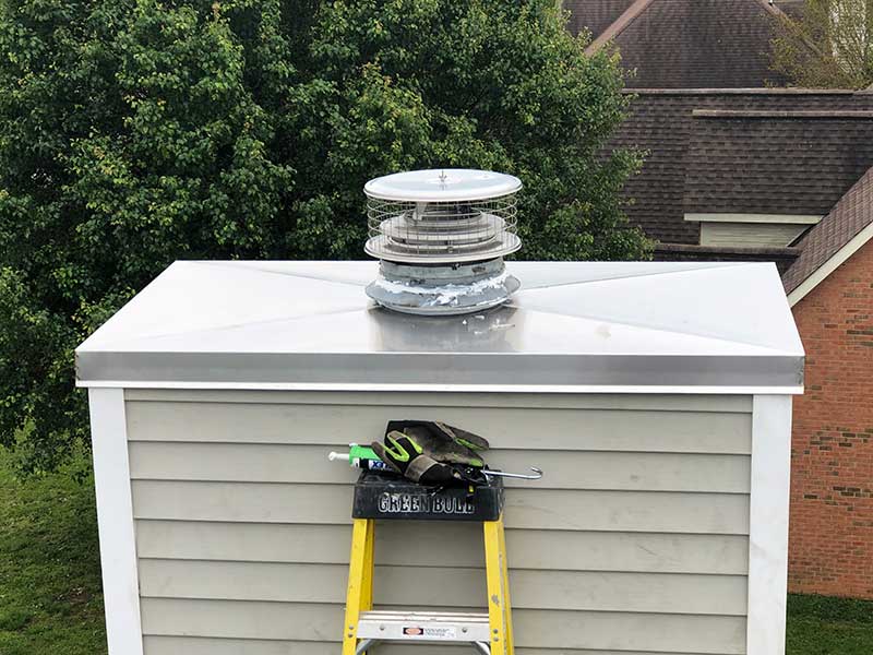Stock photo of stainless steel prefab crown - Sweeps and Ladders Prefab stainless steel crown with cap on top and ladder leaning against chimney. Homes and trees in the background.