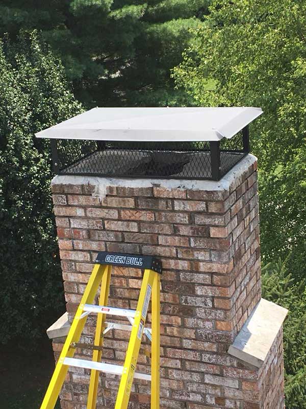 New rectangular chimney cap on brick chimney with trees in the background and ladder leaning against chimney.