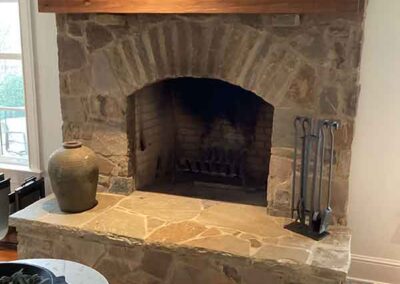 Arched fireplace made of stone with large wood mantel. Crock sitting on hearth along with tools.