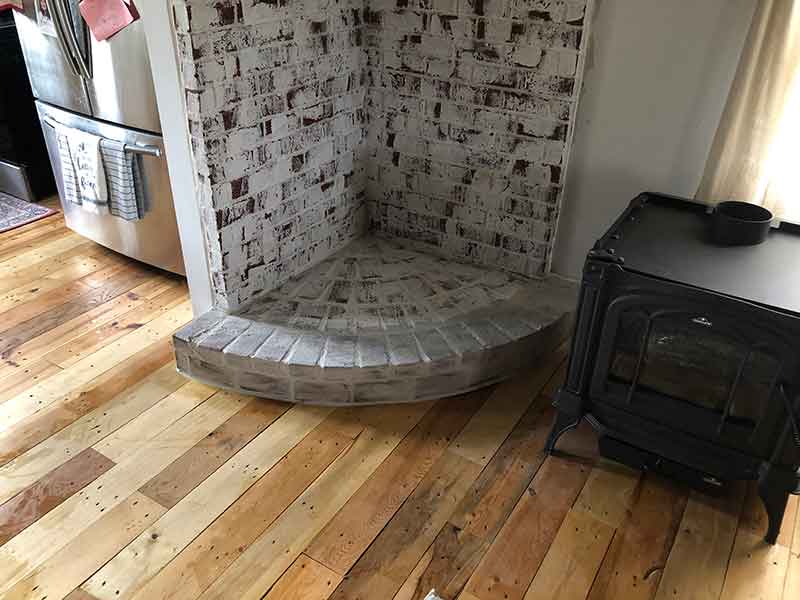 Corner brick masonry fan shaped hearth with free-standing stove to the right and refrigerator to the left.