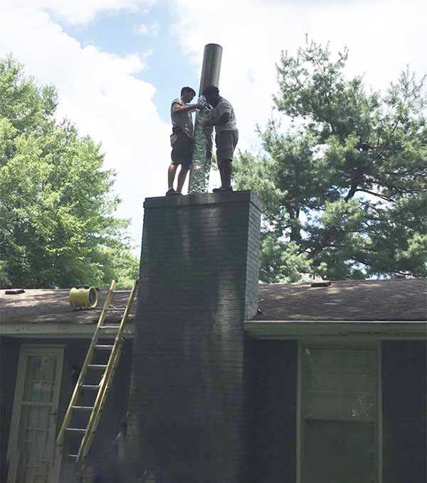 Two techs feeding stainless steel into chimney, ladder on side of house and trees in the background.