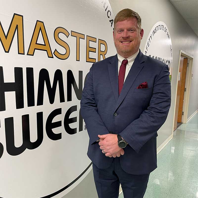 Tommy Nelms Owner - Sweeps and Ladders Tommy Nelms - Owner dressed in suit in front of Master Chimney Sweep sign.