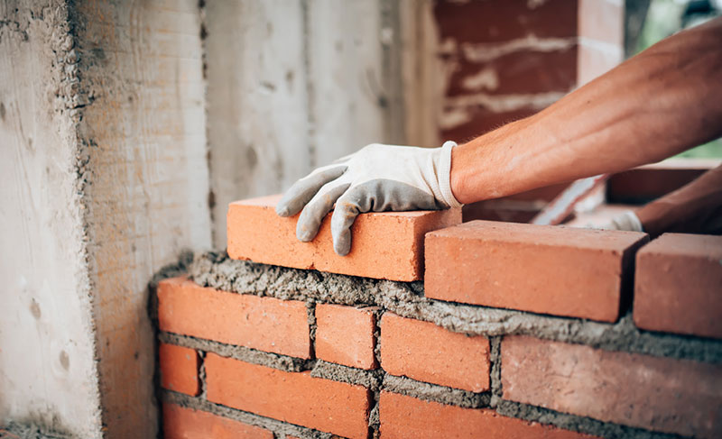Gloved hand laying brick on fresh mortar and stack of bricks.