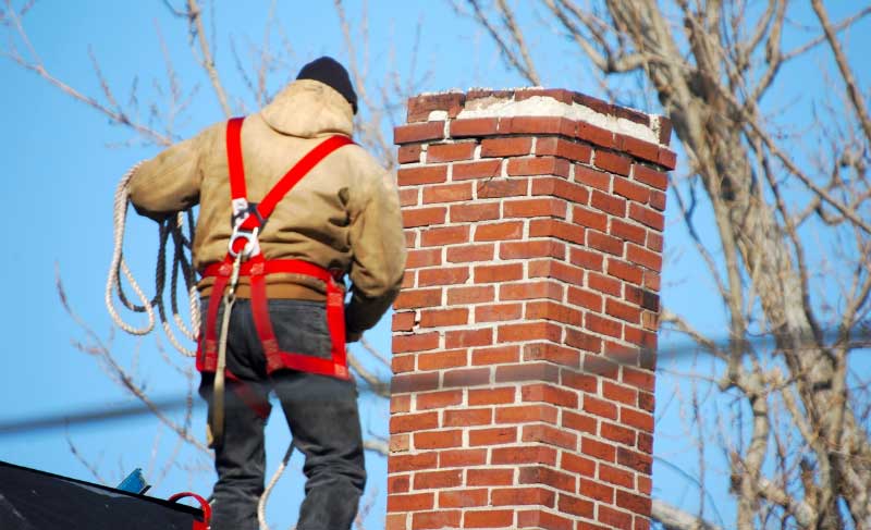 Man wearing a red safety harness on a roof holding rope next to chimney